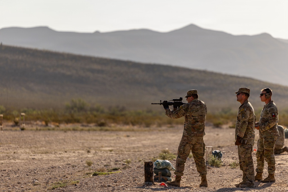 Three military personnel in camouflage uniforms are conducting a weapons qualification or training exercise at an outdoor shooting range in a desert environment. One soldier is in a standing firing position aiming a rifle downrange, while two others observe or supervise. The backdrop features arid terrain with sparse vegetation and layered mountain ridgelines in the distance.