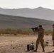 Three military personnel in camouflage uniforms are conducting a weapons qualification or training exercise at an outdoor shooting range in a desert environment. One soldier is in a standing firing position aiming a rifle downrange, while two others observe or supervise. The backdrop features arid terrain with sparse vegetation and layered mountain ridgelines in the distance.
