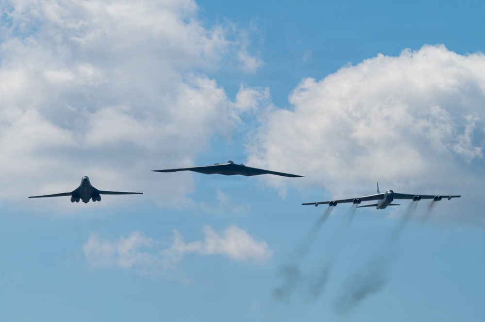 Three distinct US Air Force strategic bomber aircraft are flying in formation against a partly cloudy blue sky. The formation includes all three of the US Air Force's bomber types flying together, a rare and notable display of airpower. The rightmost aircraft is producing visible engine exhaust trails.