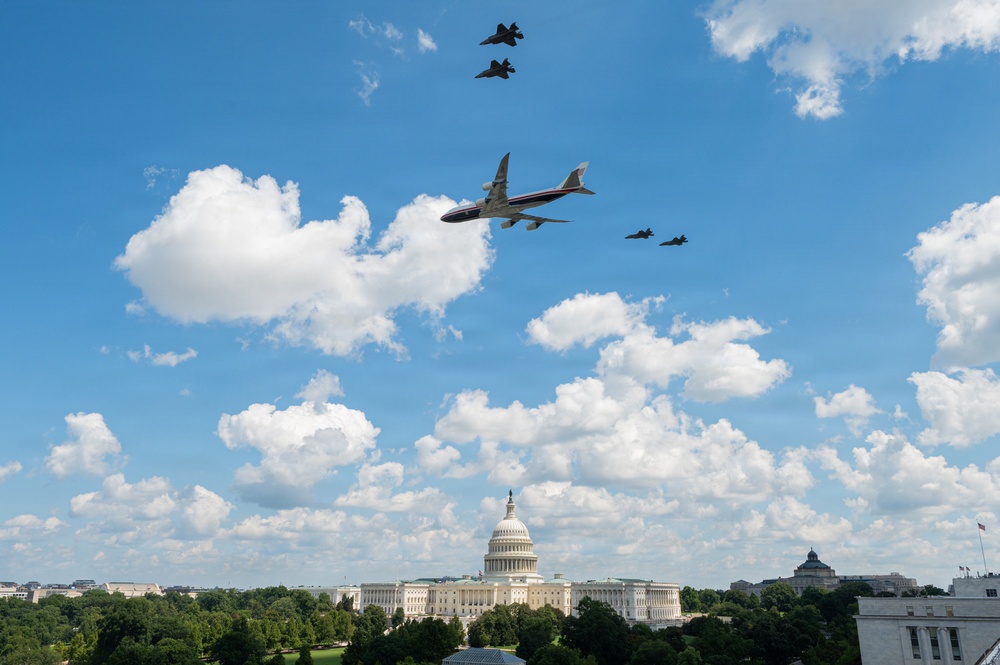 A large commercial-style aircraft with a distinctive blue, white, and red livery consistent with Air Force One flies over Washington D.C., escorted by four military fighter jets. The U.S. Capitol Building is prominently visible in the background below. The scene appears to be a ceremonial flyover captured on a partly cloudy day.