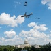 A large commercial-style aircraft with a distinctive blue, white, and red livery consistent with Air Force One flies over Washington D.C., escorted by four military fighter jets. The U.S. Capitol Building is prominently visible in the background below. The scene appears to be a ceremonial flyover captured on a partly cloudy day.
