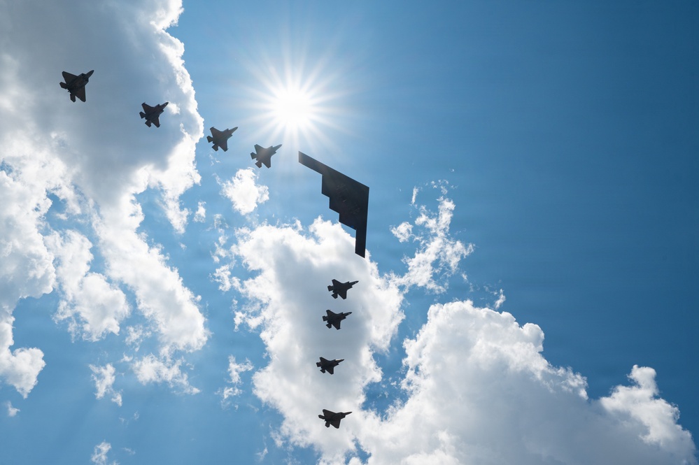A formation of military aircraft flies in silhouette against a bright blue sky with large cumulus clouds. The formation includes one large flying-wing stealth bomber (consistent with a B-2 Spirit) flanked and trailed by multiple smaller fighter jets (consistent with F-22 Raptors or F-35s). The sun is positioned directly behind the formation, creating a dramatic starburst backlit effect.