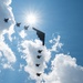 A formation of military aircraft flies in silhouette against a bright blue sky with large cumulus clouds. The formation includes one large flying-wing stealth bomber (consistent with a B-2 Spirit) flanked and trailed by multiple smaller fighter jets (consistent with F-22 Raptors or F-35s). The sun is positioned directly behind the formation, creating a dramatic starburst backlit effect.