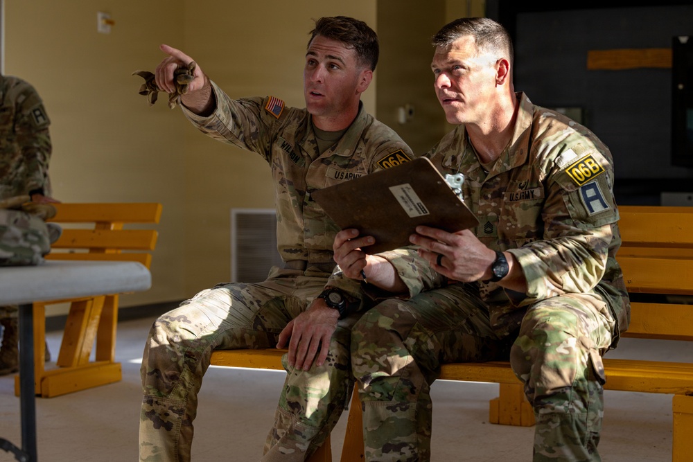Two U.S. Army soldiers in OCP (Operational Camouflage Pattern) uniforms are seated on a yellow wooden bench, reviewing a document together. One soldier, identified by a name tape reading 'WILSON,' is pointing in a direction while the other holds what appears to be a manila folder or document. Both wear unit patches indicating '06A' and '06B' designations along with the U.S. Army patch.
