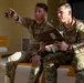 Two U.S. Army soldiers in OCP (Operational Camouflage Pattern) uniforms are seated on a yellow wooden bench, reviewing a document together. One soldier, identified by a name tape reading 'WILSON,' is pointing in a direction while the other holds what appears to be a manila folder or document. Both wear unit patches indicating '06A' and '06B' designations along with the U.S. Army patch.