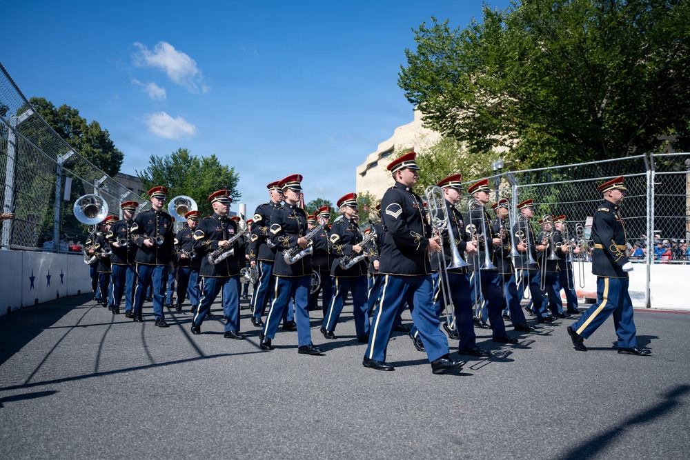 A military marching band is parading along a paved street, dressed in formal dark uniforms with red-plumed caps and gold trim. The musicians are carrying a variety of brass and woodwind instruments while marching in formation. The event appears to take place outdoors on a clear, sunny day near a fenced parade route.
