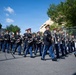A military marching band is parading along a paved street, dressed in formal dark uniforms with red-plumed caps and gold trim. The musicians are carrying a variety of brass and woodwind instruments while marching in formation. The event appears to take place outdoors on a clear, sunny day near a fenced parade route.