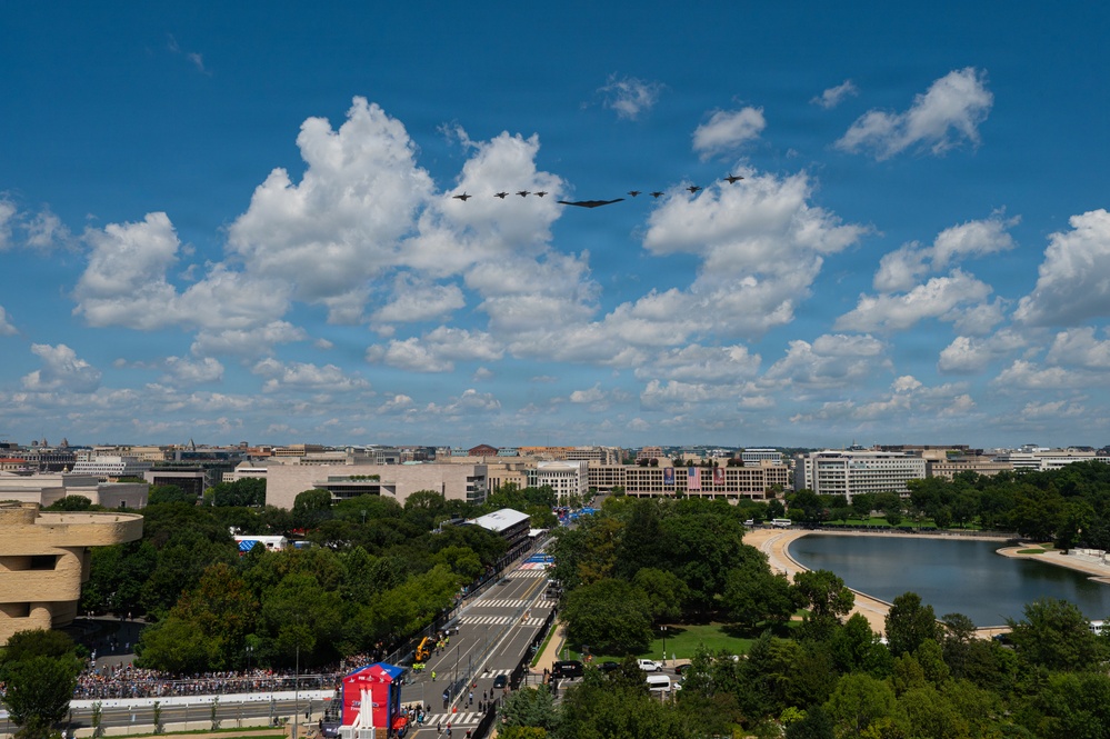 An aerial flyover formation of military aircraft, including what appears to be a B-2 Spirit stealth bomber flanked by fighter jets, passes over Washington D.C. during a public event. The image is taken from an elevated vantage point showing the National Mall area with a large crowd gathered along a boulevard. A reflecting pool or tidal basin is visible to the right, with the city's federal buildings stretching toward the horizon.