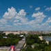An aerial flyover formation of military aircraft, including what appears to be a B-2 Spirit stealth bomber flanked by fighter jets, passes over Washington D.C. during a public event. The image is taken from an elevated vantage point showing the National Mall area with a large crowd gathered along a boulevard. A reflecting pool or tidal basin is visible to the right, with the city's federal buildings stretching toward the horizon.