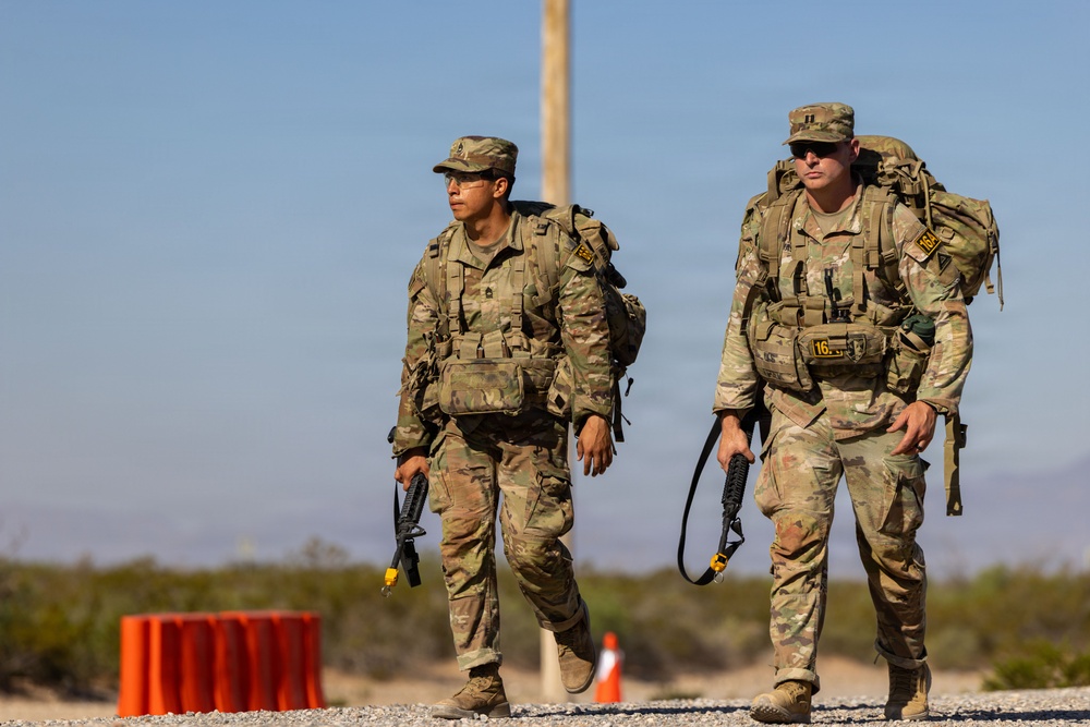 Two U.S. Army soldiers in Operational Camouflage Pattern (OCP) uniforms are conducting what appears to be a ruck march in a desert environment. Both soldiers are carrying large rucksacks and wearing tactical vests with equipment. The setting appears to be a training area or military installation based on the road, orange traffic cones, and utility pole visible in the background.