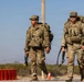 Two U.S. Army soldiers in Operational Camouflage Pattern (OCP) uniforms are conducting what appears to be a ruck march in a desert environment. Both soldiers are carrying large rucksacks and wearing tactical vests with equipment. The setting appears to be a training area or military installation based on the road, orange traffic cones, and utility pole visible in the background.