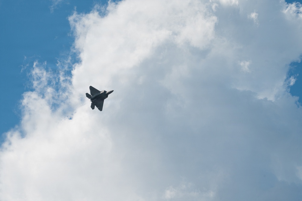 A single military jet aircraft is photographed in flight against a partly cloudy blue sky. The aircraft appears to be a fifth-generation stealth fighter, consistent with an F-22 Raptor silhouette, captured from a ground-level upward angle. The image shows the aircraft in a banking maneuver, likely during an airshow or training flight.