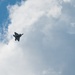 A single military jet aircraft is photographed in flight against a partly cloudy blue sky. The aircraft appears to be a fifth-generation stealth fighter, consistent with an F-22 Raptor silhouette, captured from a ground-level upward angle. The image shows the aircraft in a banking maneuver, likely during an airshow or training flight.