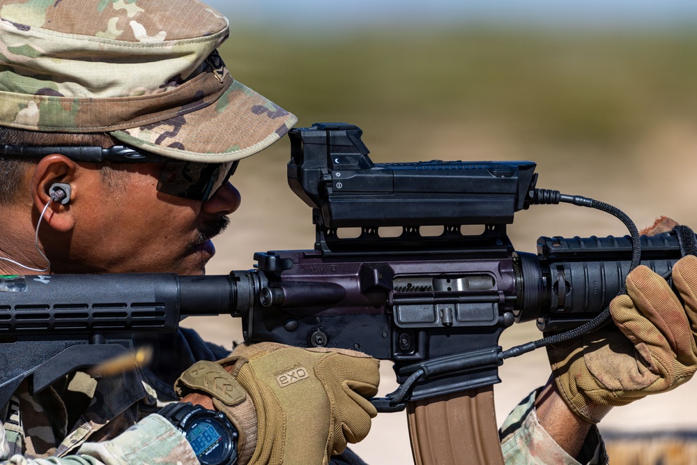 A soldier in MultiCam/OCP camouflage uniform is aiming an M4/M16-style carbine equipped with an advanced optic sight system. The image is a close-up shot capturing fine details of both the shooter and the weapon system. A spent cartridge casing appears to be ejecting from the weapon at the lower left of the frame.