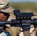 A soldier in MultiCam/OCP camouflage uniform is aiming an M4/M16-style carbine equipped with an advanced optic sight system. The image is a close-up shot capturing fine details of both the shooter and the weapon system. A spent cartridge casing appears to be ejecting from the weapon at the lower left of the frame.