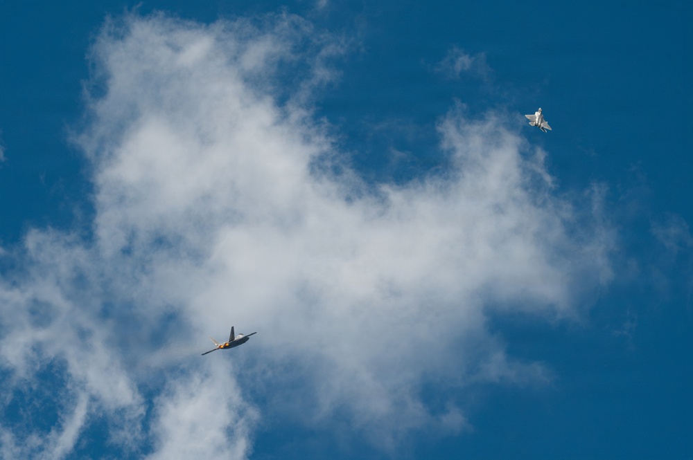 Two military jet aircraft are captured in flight against a blue sky with scattered white clouds. The aircraft appear to be flying in proximity to one another, possibly during an airshow or training exercise. The lower aircraft displays visible orange/yellow coloring on its underside, while the upper aircraft appears in a lighter gray tone.