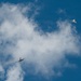 Two military jet aircraft are captured in flight against a blue sky with scattered white clouds. The aircraft appear to be flying in proximity to one another, possibly during an airshow or training exercise. The lower aircraft displays visible orange/yellow coloring on its underside, while the upper aircraft appears in a lighter gray tone.