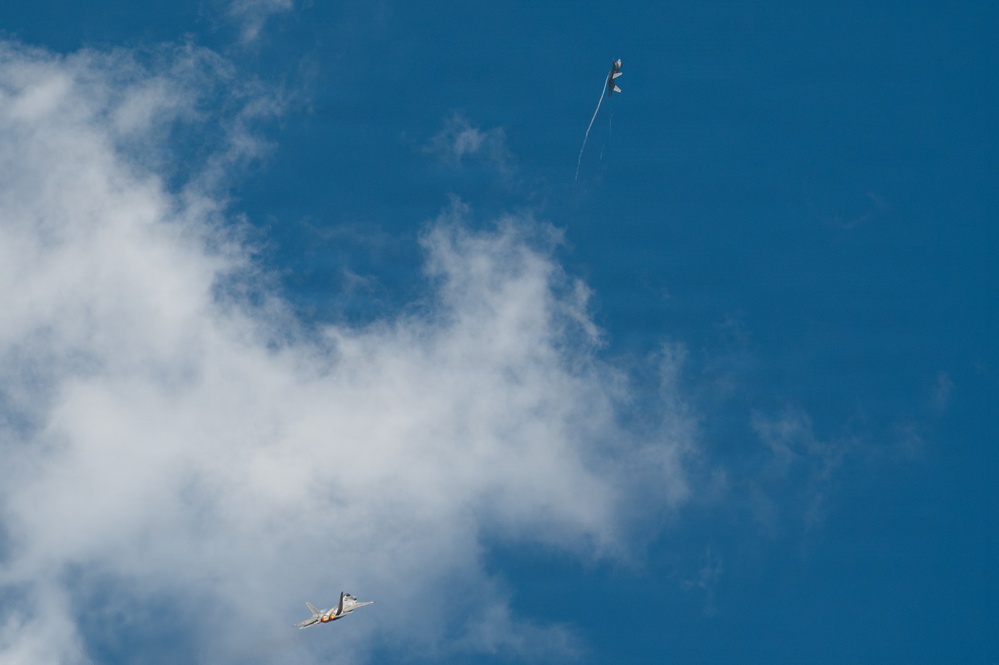 The image shows a military jet aircraft flying at low altitude against a partly cloudy blue sky. Above the jet, a smaller object — appearing to be a target drone or towed aerial target — is visible with a trailing streamer or tow line. This appears to capture a live-fire or intercept training exercise.