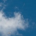 The image shows a military jet aircraft flying at low altitude against a partly cloudy blue sky. Above the jet, a smaller object — appearing to be a target drone or towed aerial target — is visible with a trailing streamer or tow line. This appears to capture a live-fire or intercept training exercise.