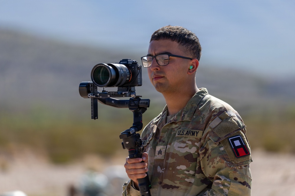 A U.S. Army Sergeant in OCP (Operational Camouflage Pattern) uniform is operating a Sony Alpha mirrorless camera mounted on a DJI gimbal stabilizer in an outdoor desert environment. The soldier appears to be performing official military documentation or media production duties. The patch on the right shoulder suggests a unit or state affiliation.