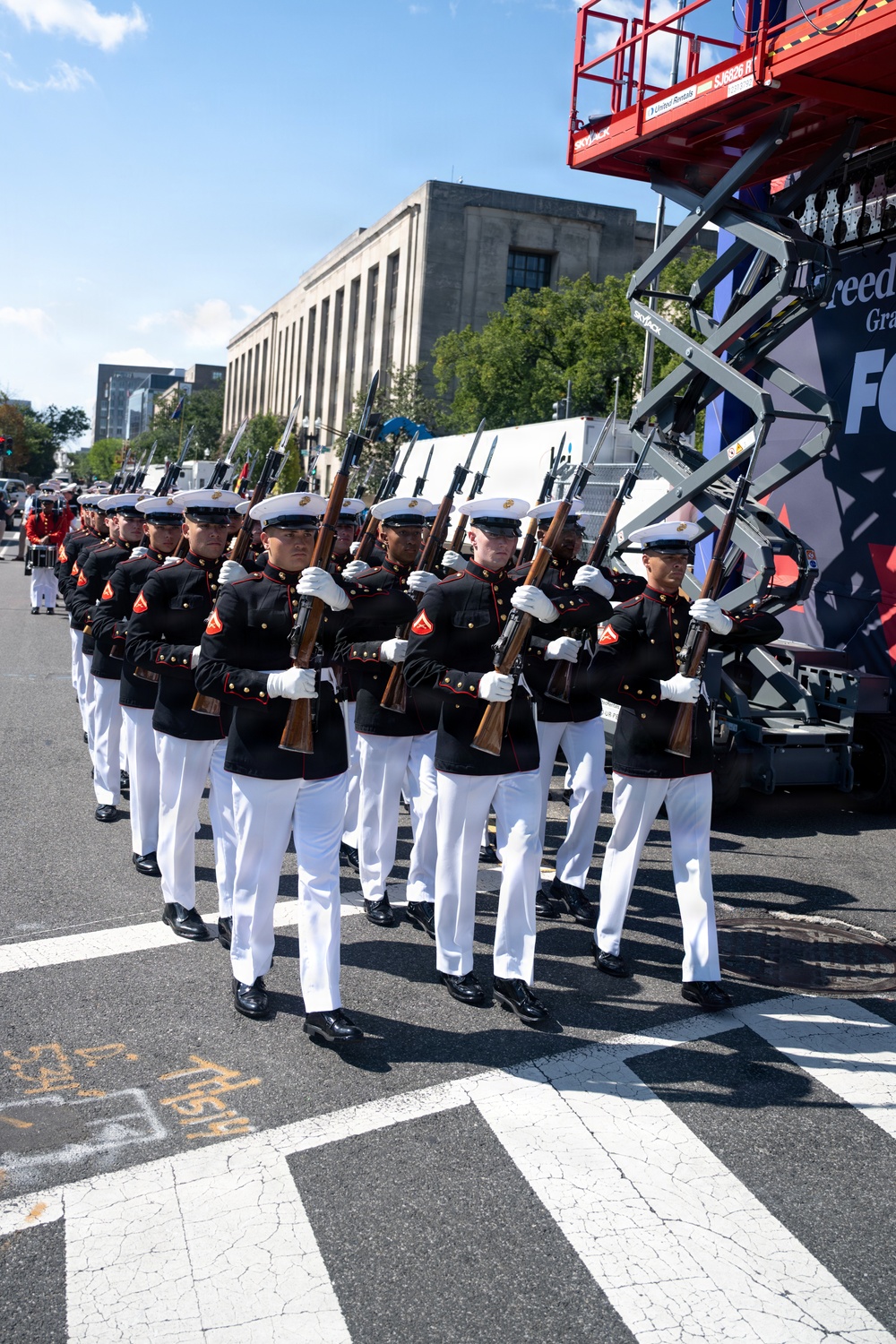 A formation of United States Marine Corps personnel marches in a parade along a city street. The Marines are dressed in their distinctive dress blues uniform with white trousers, white gloves, and white covers (caps), carrying rifles with bayonets fixed. A scissor lift with red framing and partial signage reading 'Freedom' is visible to the right, suggesting a formal public ceremony or patriotic event.