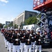 A formation of United States Marine Corps personnel marches in a parade along a city street. The Marines are dressed in their distinctive dress blues uniform with white trousers, white gloves, and white covers (caps), carrying rifles with bayonets fixed. A scissor lift with red framing and partial signage reading 'Freedom' is visible to the right, suggesting a formal public ceremony or patriotic event.