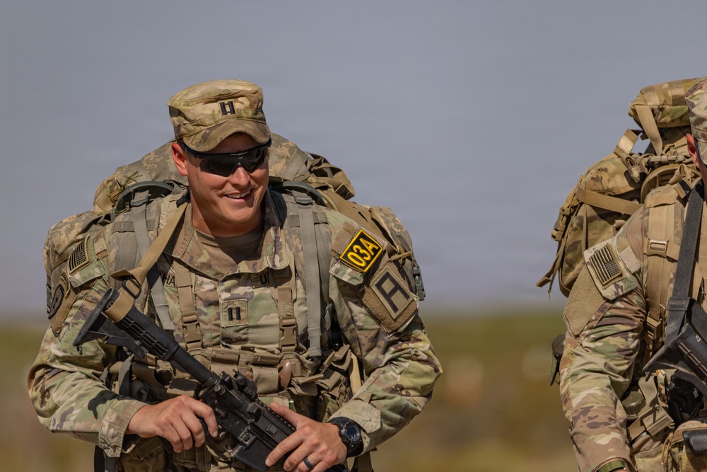 A smiling U.S. Army Captain is photographed during what appears to be a ruck march or field training exercise in an arid environment. He is wearing full combat gear including a rucksack, body armor, and carrying an assault rifle. A second soldier is partially visible to the right.