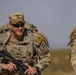 A smiling U.S. Army Captain is photographed during what appears to be a ruck march or field training exercise in an arid environment. He is wearing full combat gear including a rucksack, body armor, and carrying an assault rifle. A second soldier is partially visible to the right.