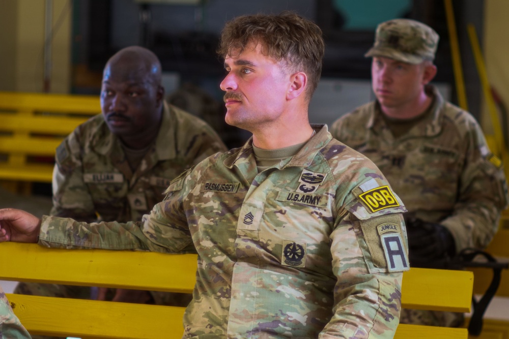 Three U.S. Army soldiers in Operational Camouflage Pattern (OCP) uniforms are seated on yellow benches, appearing to attend a briefing or formation. The central soldier, identified by name tape as 'RASMUSSEN,' wears a Staff Sergeant rank insignia, a '09B' unit patch, a 'Jungle' tab, and an 'A' (Ranger) tab on his left shoulder. The soldiers appear attentive and are in an indoor military facility.
