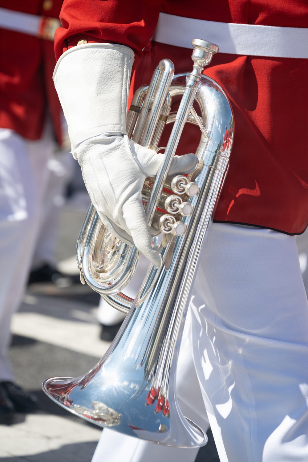 A close-up photograph of a marching band musician holding a silver flugelhorn or mellophone at their side. The performer is dressed in a red uniform jacket with white trousers and white leather gloves, consistent with a military or ceremonial marching band. The image is tightly cropped, focusing on the instrument and the gloved hand holding it.