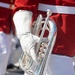 A close-up photograph of a marching band musician holding a silver flugelhorn or mellophone at their side. The performer is dressed in a red uniform jacket with white trousers and white leather gloves, consistent with a military or ceremonial marching band. The image is tightly cropped, focusing on the instrument and the gloved hand holding it.