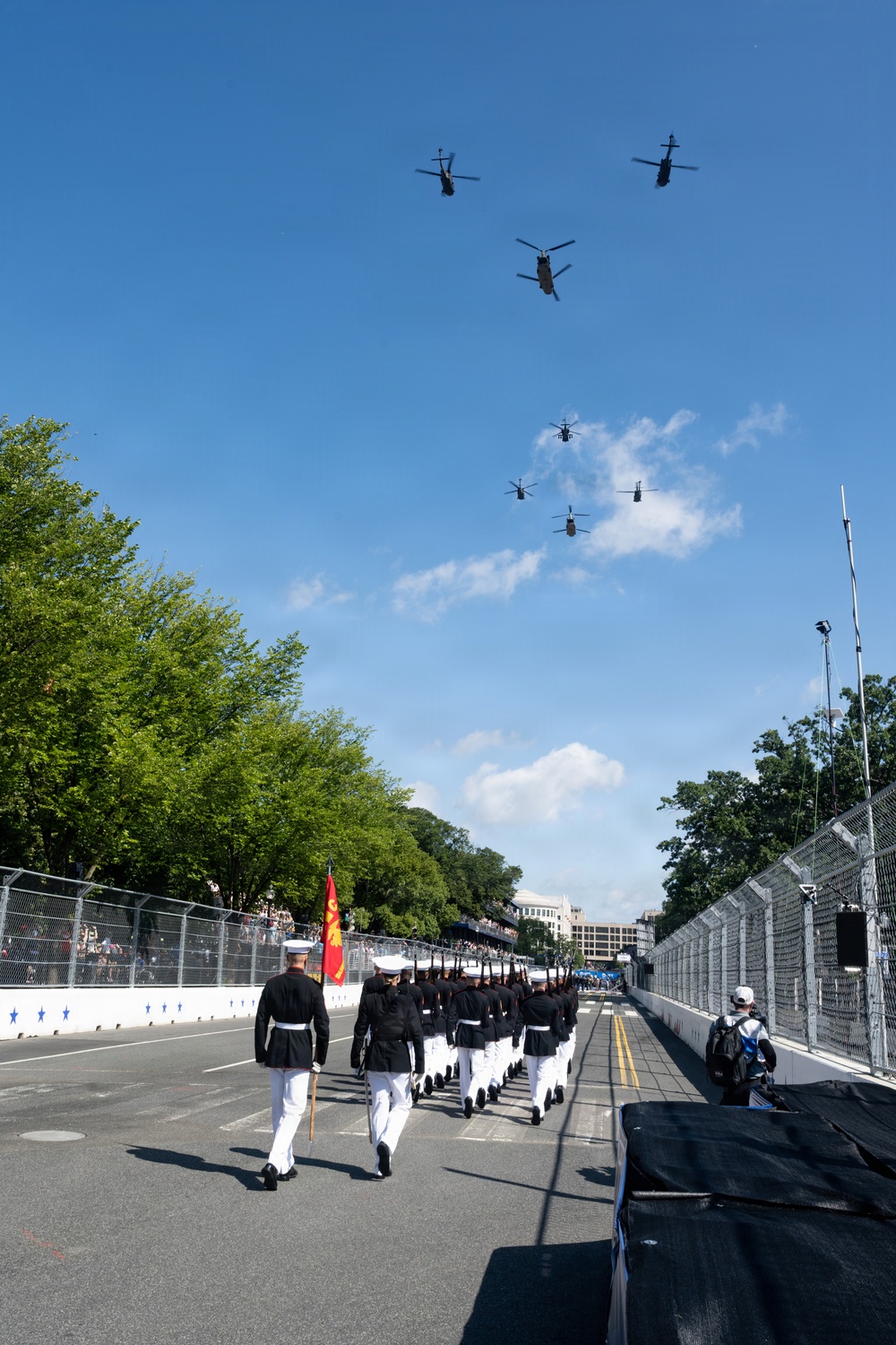 A military parade is taking place on a closed street, with a formation of uniformed personnel marching in black jackets and white trousers, led by an officer carrying a ceremonial sword. A red unit guidon flag is visible near the front of the formation. Overhead, seven military helicopters fly in a formation pattern against a clear blue sky.