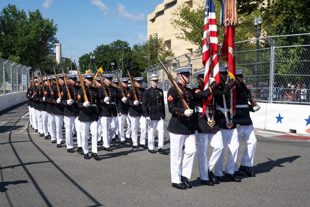 A U.S. Marine Corps ceremonial unit is marching in formation along a city street, carrying rifles at shoulder arms and bearing color guard flags including the American flag. The Marines are dressed in their distinctive dress blues uniform with white trousers, white gloves, and white peaked caps. The event appears to be a formal parade taking place in an urban setting with spectators visible behind security fencing.