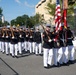 A U.S. Marine Corps ceremonial unit is marching in formation along a city street, carrying rifles at shoulder arms and bearing color guard flags including the American flag. The Marines are dressed in their distinctive dress blues uniform with white trousers, white gloves, and white peaked caps. The event appears to be a formal parade taking place in an urban setting with spectators visible behind security fencing.
