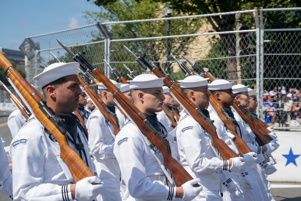 A formation of U.S. Navy Ceremonial Guard members marches in a parade, dressed in white dress uniforms and white sailor caps. They are carrying rifles with fixed bayonets at a uniform angle, suggesting a precision drill or honor guard performance. Spectators and security fencing are visible in the background.