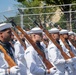 A formation of U.S. Navy Ceremonial Guard members marches in a parade, dressed in white dress uniforms and white sailor caps. They are carrying rifles with fixed bayonets at a uniform angle, suggesting a precision drill or honor guard performance. Spectators and security fencing are visible in the background.