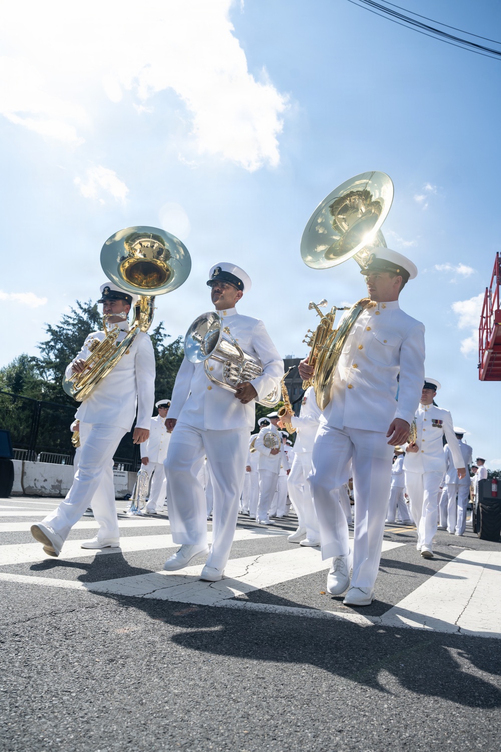 A military marching band in white dress uniforms is parading along a paved street under a partly cloudy sky. The musicians are carrying brass instruments including sousaphones and a French horn, marching in formation. The scene appears to be a formal military ceremony or public parade event.