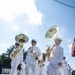 A military marching band in white dress uniforms is parading along a paved street under a partly cloudy sky. The musicians are carrying brass instruments including sousaphones and a French horn, marching in formation. The scene appears to be a formal military ceremony or public parade event.