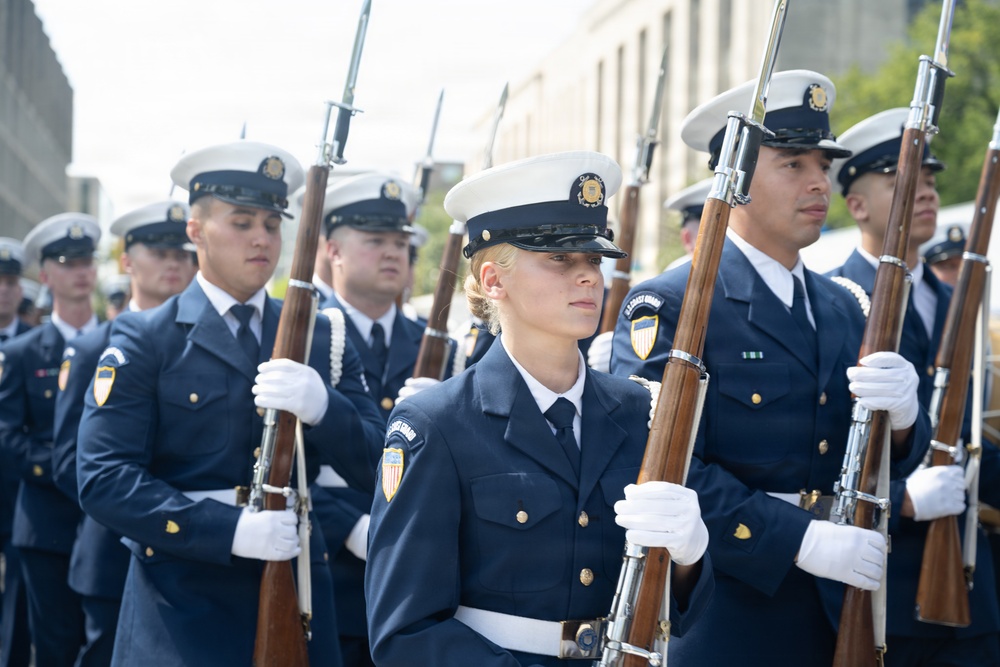 A formation of U.S. Coast Guard personnel marching in a ceremonial parade or formal procession. The members are in dress uniform carrying rifles with bayonets fixed, wearing white gloves and white-topped service caps. The scene appears to take place on an urban street with buildings visible in the background.