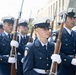 A formation of U.S. Coast Guard personnel marching in a ceremonial parade or formal procession. The members are in dress uniform carrying rifles with bayonets fixed, wearing white gloves and white-topped service caps. The scene appears to take place on an urban street with buildings visible in the background.