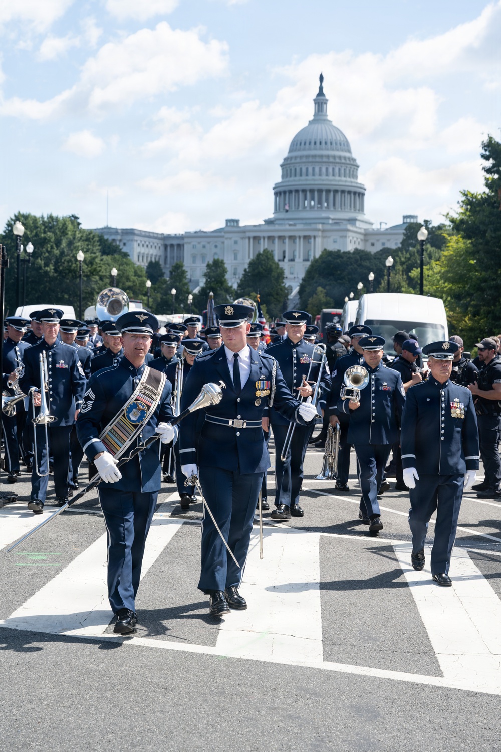 A U.S. Air Force military marching band is parading down a street in Washington, D.C., with the United States Capitol Building prominently visible in the background. Personnel are dressed in formal dark blue Air Force dress uniforms, with several members carrying instruments and a drum major leading with a ceremonial mace. The event appears to be a formal military parade or ceremonial procession along the National Mall area.