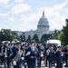 A U.S. Air Force military marching band is parading down a street in Washington, D.C., with the United States Capitol Building prominently visible in the background. Personnel are dressed in formal dark blue Air Force dress uniforms, with several members carrying instruments and a drum major leading with a ceremonial mace. The event appears to be a formal military parade or ceremonial procession along the National Mall area.
