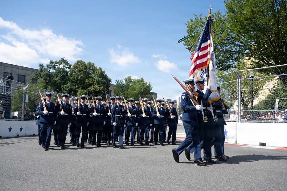 A military color guard and drill unit marches in formation during an outdoor parade or ceremonial event. Personnel are dressed in dark blue dress uniforms with white covers (caps) and white gloves, carrying rifles at a uniform angle. The lead color guard bearers carry the United States flag and at least one additional organizational flag.