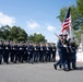 A military color guard and drill unit marches in formation during an outdoor parade or ceremonial event. Personnel are dressed in dark blue dress uniforms with white covers (caps) and white gloves, carrying rifles at a uniform angle. The lead color guard bearers carry the United States flag and at least one additional organizational flag.