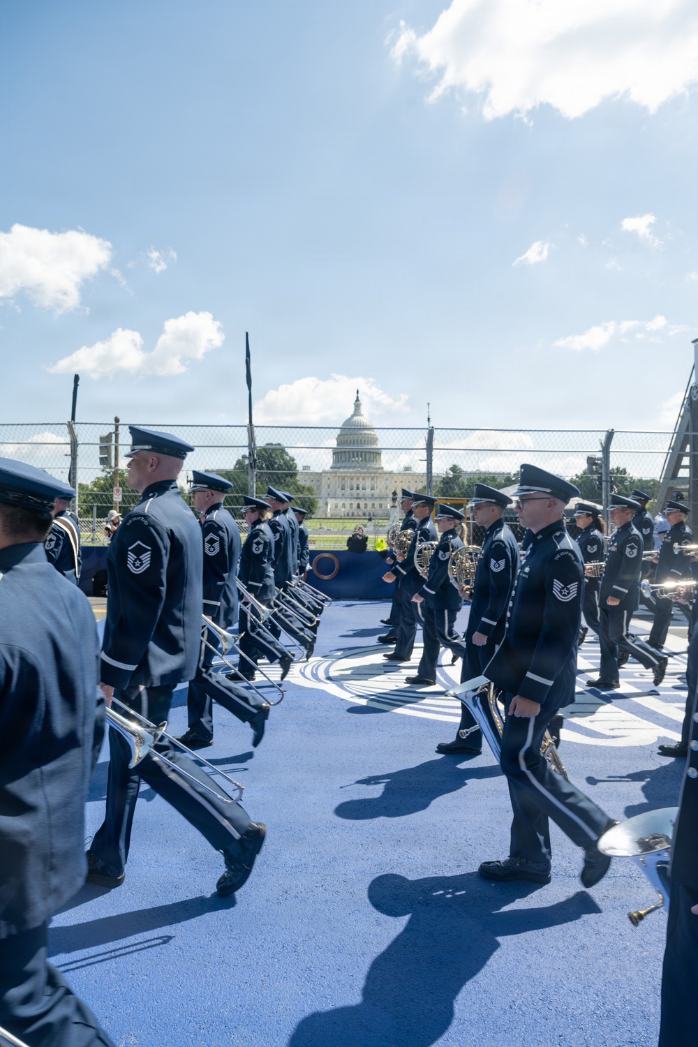 A military marching band is performing or rehearsing on a blue-painted outdoor stage or platform near the U.S. Capitol building in Washington, D.C. The musicians are dressed in dark navy blue dress uniforms with white-trimmed caps and are carrying brass instruments including trombones and French horns. The event appears to be an official ceremonial performance given the formal attire and prominent location.