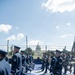 A military marching band is performing or rehearsing on a blue-painted outdoor stage or platform near the U.S. Capitol building in Washington, D.C. The musicians are dressed in dark navy blue dress uniforms with white-trimmed caps and are carrying brass instruments including trombones and French horns. The event appears to be an official ceremonial performance given the formal attire and prominent location.