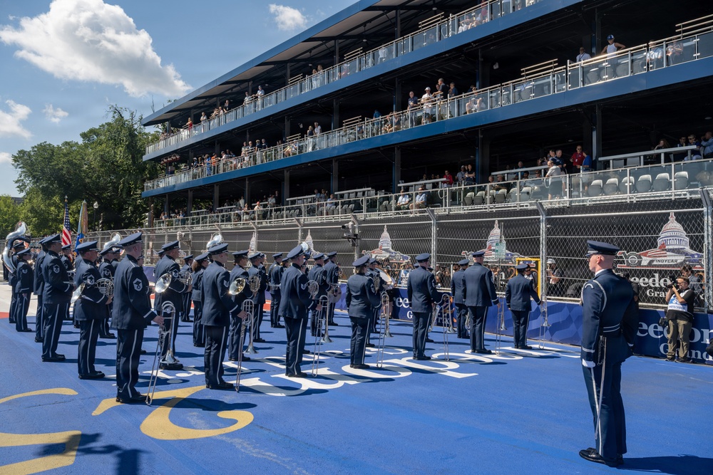 A military band, likely the United States Air Force Band, is performing on a blue surface at what appears to be the IndyCar Grand Prix on the streets of Washington D.C. The musicians are dressed in formal dark blue uniforms with white caps and are playing brass instruments including trombones and French horns. A multi-tiered grandstand filled with spectators is visible in the background, along with a depiction of the U.S. Capitol building on signage.