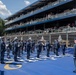 A military band, likely the United States Air Force Band, is performing on a blue surface at what appears to be the IndyCar Grand Prix on the streets of Washington D.C. The musicians are dressed in formal dark blue uniforms with white caps and are playing brass instruments including trombones and French horns. A multi-tiered grandstand filled with spectators is visible in the background, along with a depiction of the U.S. Capitol building on signage.