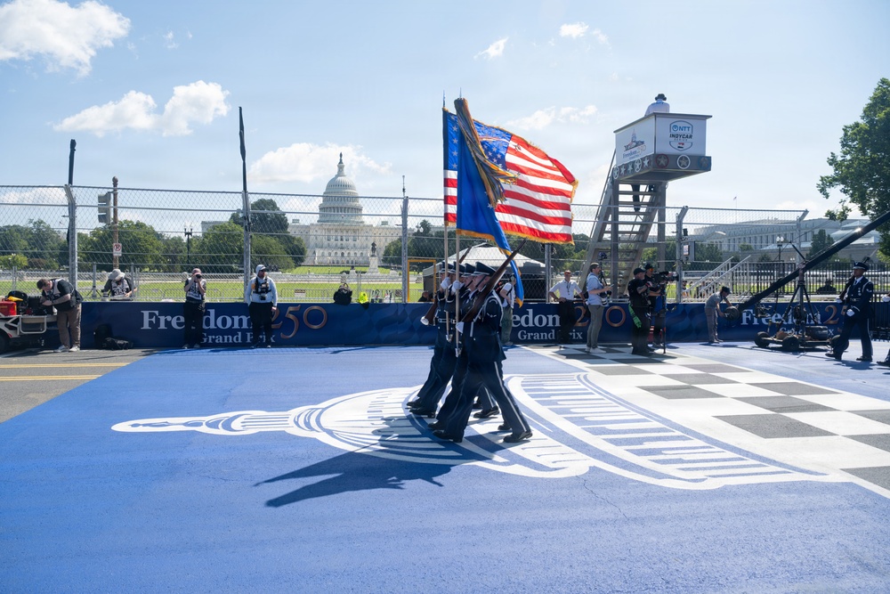 A military color guard ceremony is taking place at the 'Freedom 250 Grand Prix,' an IndyCar racing event held near the U.S. Capitol in Washington, D.C. Uniformed service members are carrying the American flag and another flag across a blue-painted start/finish area featuring a checkered pattern and decorative logo. The event appears to be a pre-race ceremony combining patriotic and motorsport elements.