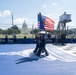 A military color guard ceremony is taking place at the 'Freedom 250 Grand Prix,' an IndyCar racing event held near the U.S. Capitol in Washington, D.C. Uniformed service members are carrying the American flag and another flag across a blue-painted start/finish area featuring a checkered pattern and decorative logo. The event appears to be a pre-race ceremony combining patriotic and motorsport elements.